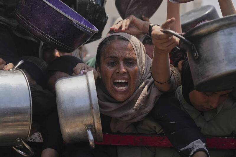 加萨民众在社区厨房排队抢领捐赠食物，一名妇女情绪激动，高举锅具呐喊求援，反映饥饿压力与援助混乱场景。（美联社）
